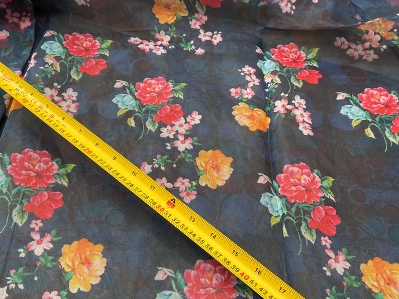 Puede incluir: Un primer plano de una tela azul oscuro transparente con estampado floral. La tela presenta flores rojas, amarillas y blancas sobre un fondo azul oscuro. Una cinta m&eacute;trica amarilla es visible en la esquina inferior derecha de la imagen.