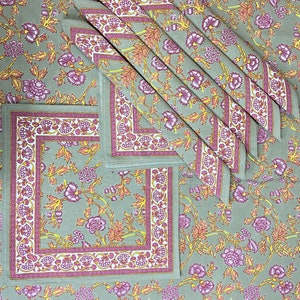 May include: A floral tablecloth and matching napkins in a sage green color. The design features purple and yellow flowers with a decorative border. The napkins are neatly folded, and the tablecloth is spread out, showcasing the pattern.