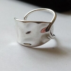 May include: A silver ring with a textured, hammered, oval-shaped band. The band is open on one side and wraps around the finger.