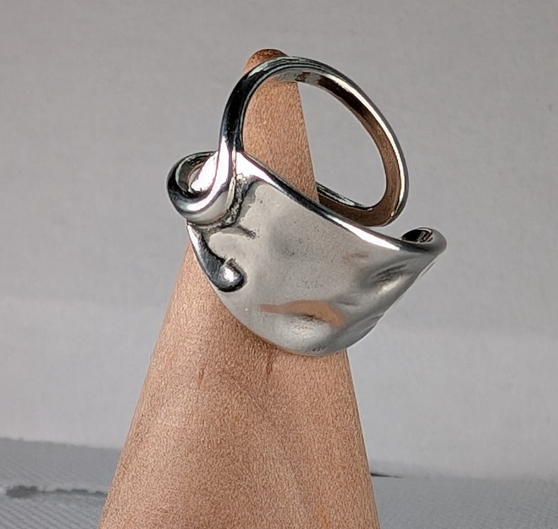 May include: A silver ring with a unique, organic, and textured design. The ring is displayed on a wooden stand.