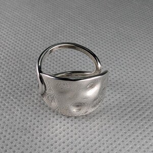 May include: A silver ring with a unique, textured, and slightly curved design. The ring is made of metal and has a smooth, polished finish.
