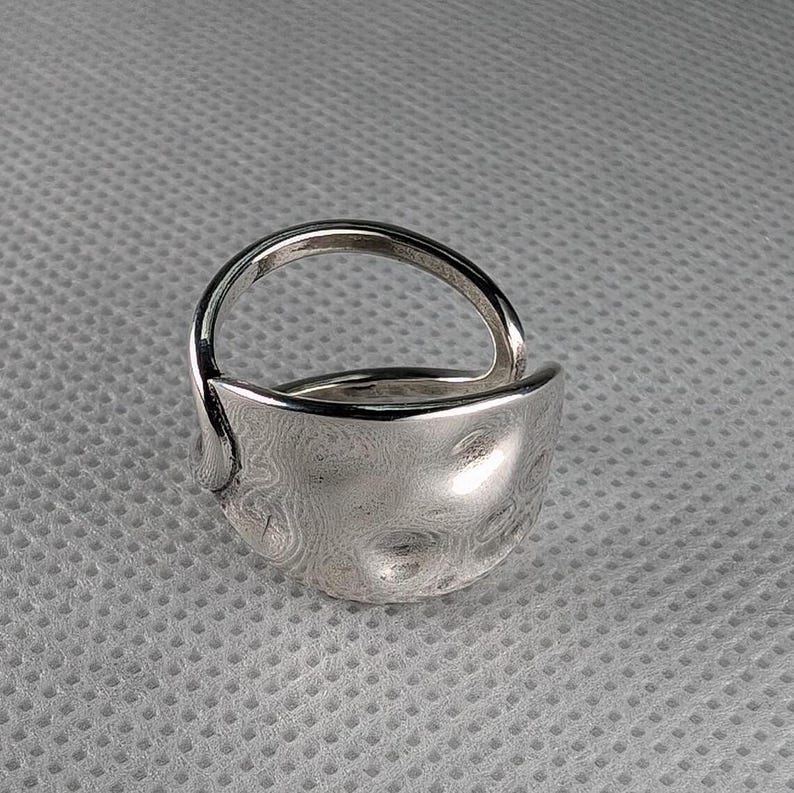 May include: A silver ring with a unique, textured, and slightly curved design. The ring is made of metal and has a smooth, polished finish.