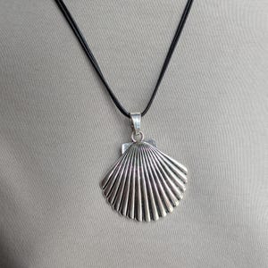 May include: A silver seashell pendant necklace. The pendant, a detailed scallop shell replica, hangs from a black cord. The necklace is shown against a neutral background.