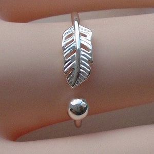 May include: A silver adjustable ring with a feather design. The ring has a small, round ball on the opposite end of the feather. The ring is shown on a person's skin.