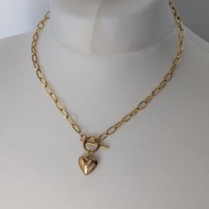 May include: Gold-coloured necklace with a heart pendant. The necklace has a chain with oval links and a toggle clasp. The heart-shaped pendant is smooth and polished, adding a touch of elegance to the jewellery.