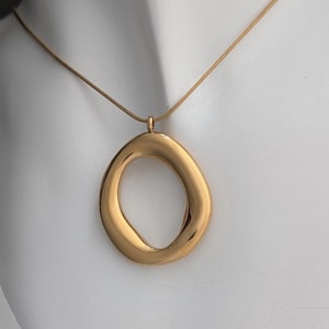 May include: A gold-coloured necklace with a large, abstract oval pendant. The pendant has a smooth, polished finish and hangs from a fine chain. The design is simple yet elegant. The necklace is shown against a white background.