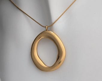 May include: A gold-toned necklace featuring a large, abstract pendant with an oval shape and a smooth, polished finish. The pendant hangs from a delicate chain, creating a simple yet elegant design. The necklace is displayed against a white background.