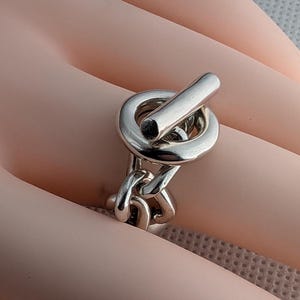 May include: A silver ring with a thick chain link design and a large, round, toggle clasp.