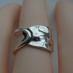 May include: A silver ring with a unique, abstract design. The ring is made of a single piece of metal that has been hammered and shaped into a curved, asymmetrical form.