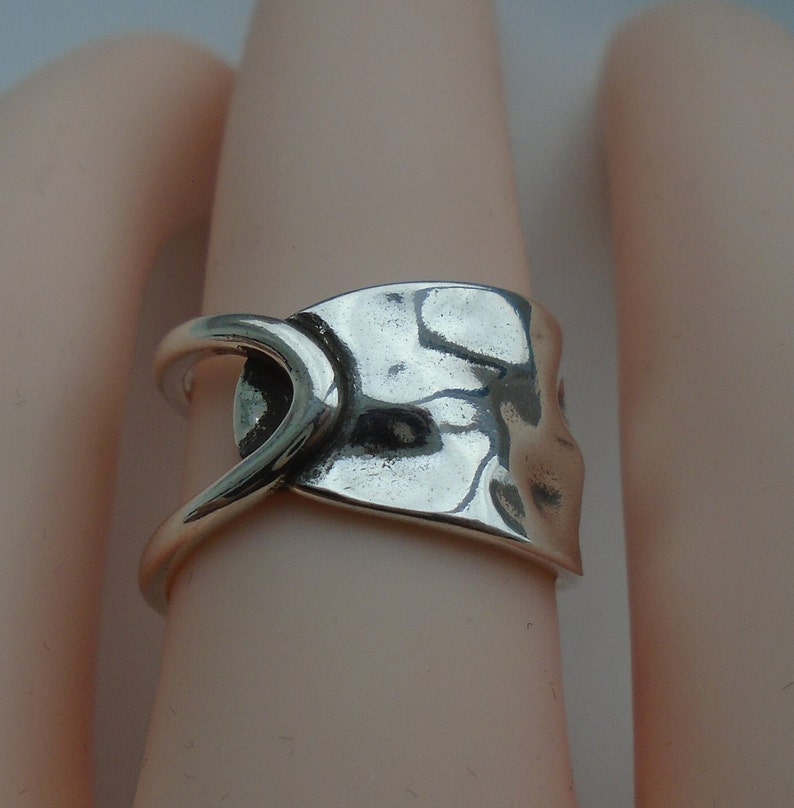 May include: A silver ring with a unique, abstract design. The ring is made of a single piece of metal that has been hammered and shaped into a curved, asymmetrical form.
