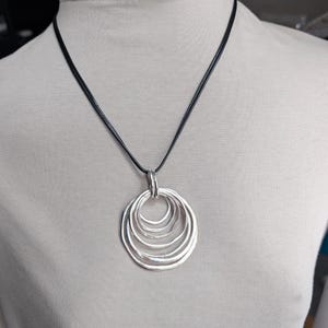 May include: Silver-toned pendant necklace featuring a series of graduated, concentric circles. The pendant hangs from a black cord. The necklace is displayed on a mannequin.