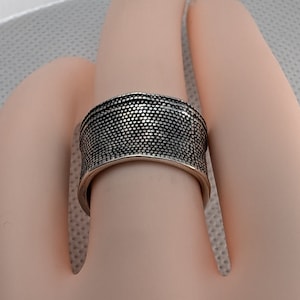 May include: A wide silver band ring with a textured surface. The ring has a hammered finish and a slightly rounded shape.