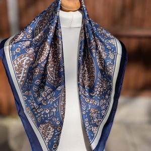 May include: A square scarf with a paisley pattern in shades of navy blue, brown, and cream. The scarf is displayed on a mannequin, showcasing its intricate floral and ornamental design. The edges are finished with a dark blue border.