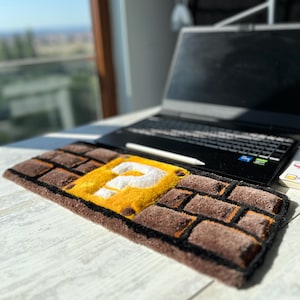 Peut inclure: Un tapis rectangulaire avec un motif de briques marron et noir, et un point d'interrogation jaune et blanc au centre. Le tapis est placé devant un ordinateur portable sur une surface blanche.