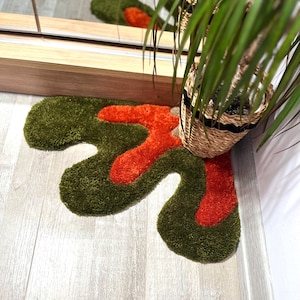 May include: A green and orange abstract shaped rug with a shaggy texture. The rug is placed in a corner of a room with a potted plant.
