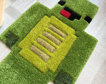 May include: A green and yellow rug in the shape of a pixelated character. The rug has a textured, plush surface with a rectangular design on the chest. The character's face features black eyes and a red mouth.