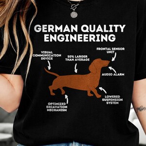 May include: Black t-shirt with the text "GERMAN QUALITY ENGINEERING" in white. A brown dachshund illustration is featured with labels such as "Visual Communication Device" and "Audio Alarm".