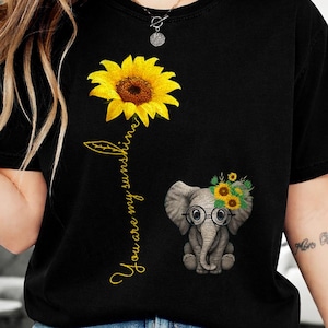 May include: Black t-shirt featuring a large yellow sunflower and a small elephant wearing glasses and a sunflower crown. The text "You are my sunshine" is written in gold script. The shirt is a casual style.