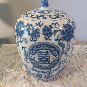 May include: A white ceramic ginger jar with a lid, featuring blue hand-painted floral patterns and a central double happiness symbol. The jar has a rounded body, a tapered neck, and a pointed finial on the lid, showcasing a classic blue and white color scheme with intricate botanical details.