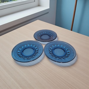 May include: Three blue glass plates with a central recessed area and a decorative pattern around the rim. The plates are arranged on a light-colored wooden surface, with one smaller plate positioned above two larger ones.