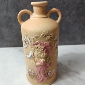 May include: A ceramic vase with a light brown glaze and a raised design of a girl holding a white bird. The girl is wearing a pink dress and a pink hat. The vase has two handles and is decorated with pink flowers.