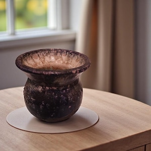 May include: A dark, textured vase with a wide, flared rim. The vase has a bulbous body and sits on a light-colored, circular coaster. The interior of the vase has a lighter, speckled appearance. The vase is placed on a wooden table.