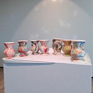May include: A collection of seven small, decorative ceramic vases. Each vase features a unique swirling pattern of colors, including red, blue, green, and brown. The vases have a bulbous shape with a narrow neck and a white rim.