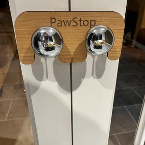 Könnte beinhalten: Ein Türstopper aus Holz mit dem eingravierten Text "PawStop". Der Türstopper hat zwei verchromte Kugeln. Der Türstopper soll verhindern, dass Türen vollständig geschlossen werden.