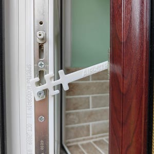 Könnte beinhalten: Ein weißer Fensterbegrenzer aus Kunststoff mit der Aufschrift AIR-STRIP, der an einem Metallfensterrahmen befestigt ist. Das Gerät verfügt über ein gekerbtes Design mit mehreren Verriegelungspositionen und ist mit sichtbaren Schrauben befestigt.