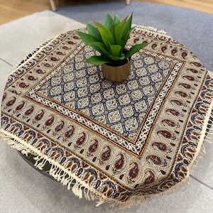 May include: A square table runner with a cream background and a blue, red, and gold paisley pattern. The runner has a fringe border and is decorated with a potted green plant.