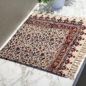 May include: A cream-colored table runner with a floral pattern in shades of red, blue, and gold. The runner has a fringed edge and is made of cotton.