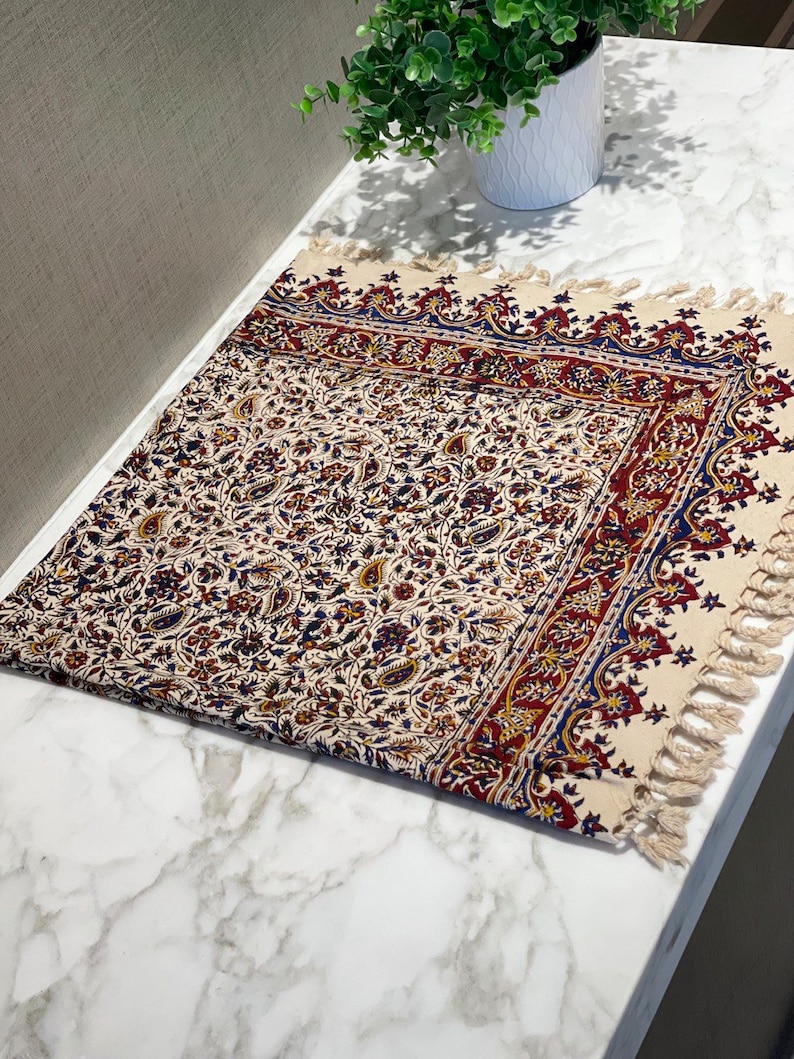 May include: A cream-colored table runner with a floral pattern in shades of red, blue, and gold. The runner has a fringed edge and is made of cotton.
