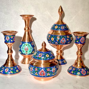 May include: A collection of copper home decor items, including two candle holders, a vase, a lidded jar, and a decorative bowl. Each piece features a blue band with floral designs in red, yellow, and white, set against a copper background.