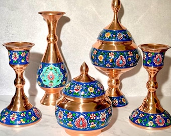 May include: A collection of copper home decor items, including two candle holders, a vase, a lidded jar, and a decorative bowl. Each piece features a blue band with floral designs in red, yellow, and white, set against a copper background.