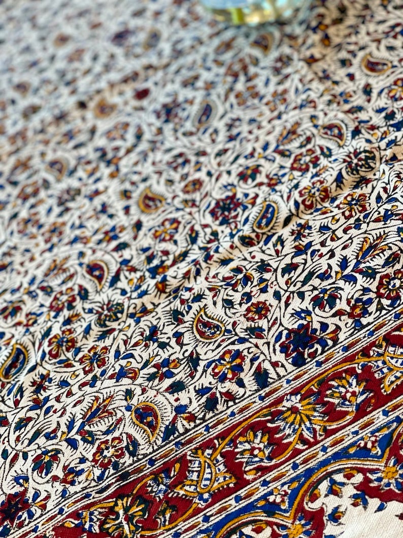 May include: A white tablecloth with a paisley and floral pattern in shades of blue, red, and yellow. The tablecloth has a red border with a paisley design.