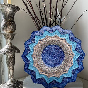 May include: A stack of decorative, layered plates with intricate blue and beige patterns. The plates have a starburst shape and are arranged on a white surface. A silver candle holder and branches with small white flowers are in the background.