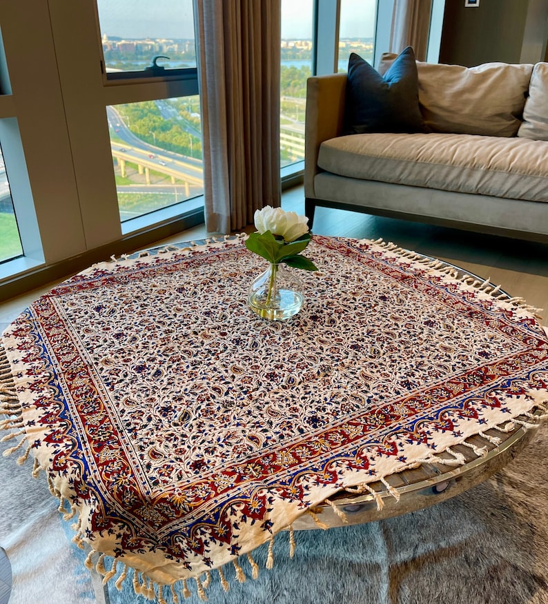 May include: A white tablecloth with a paisley pattern and fringe trim. The tablecloth is on a round table with a glass top. A vase with a white flower is in the center of the table.