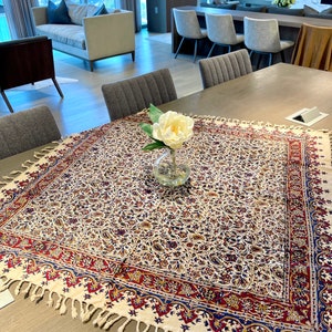 May include: A white tablecloth with a red, blue, and gold floral pattern and fringe trim. A single white flower in a clear glass vase sits in the center of the tablecloth.