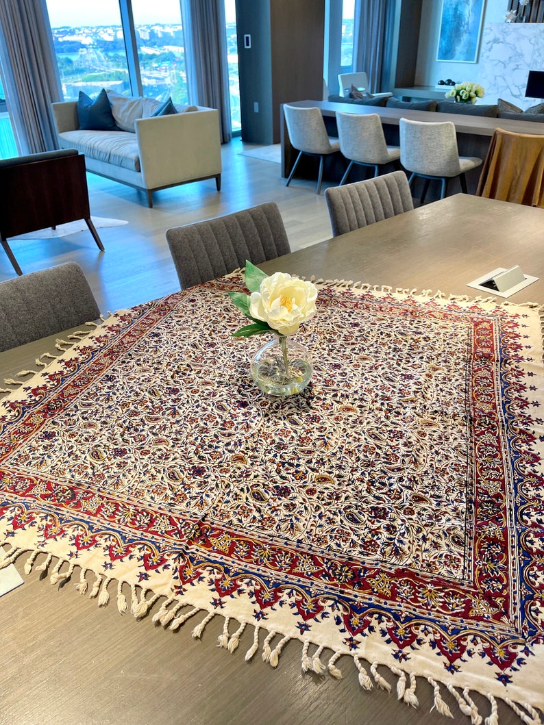 May include: A white tablecloth with a red, blue, and gold floral pattern and fringe trim. A single white flower in a clear glass vase sits in the center of the tablecloth.