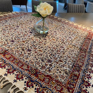 May include: A white tablecloth with a red, blue, and gold floral pattern. The tablecloth has a fringe border and is laid on a wooden table. A white flower in a clear glass vase is in the center of the table.