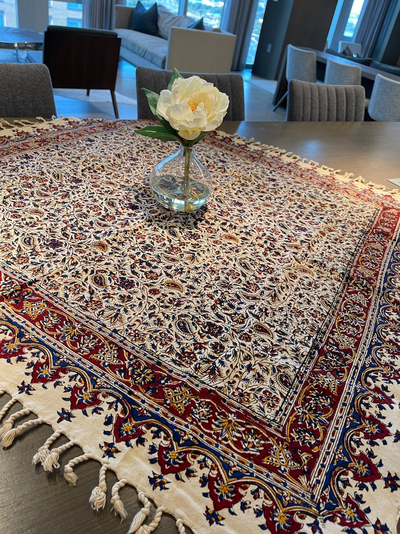 May include: A white tablecloth with a red, blue, and gold floral pattern. The tablecloth has a fringe border and is laid on a wooden table. A white flower in a clear glass vase is in the center of the table.
