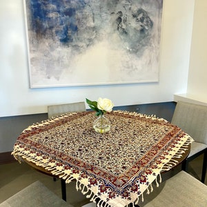 May include: A round table with a white tablecloth featuring a floral pattern in shades of red, blue, and gold. The tablecloth has a fringe border. A white flower in a glass vase sits in the center of the table.