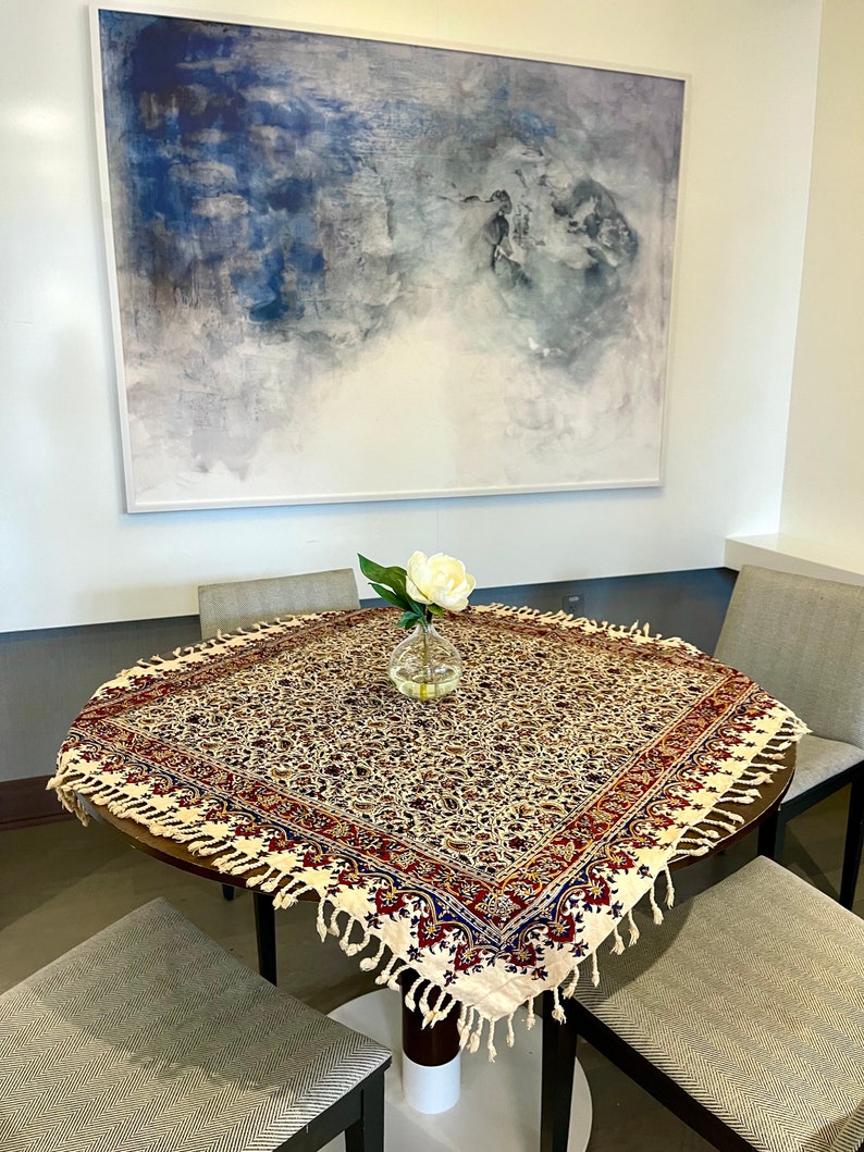 May include: A round table with a white tablecloth featuring a floral pattern in shades of red, blue, and gold. The tablecloth has a fringe border. A white flower in a glass vase sits in the center of the table.