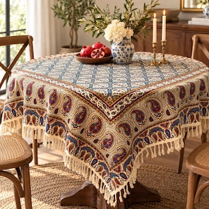May include: A square tablecloth featuring a blue and white grid pattern on top with a wide border of red, blue, and gold paisley motifs. The edges are finished with long, cream-colored fringe tassels hanging down from all four sides of the fabric.