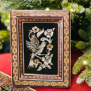 Pode incluir: Moldura retangular ornamentada com fundo preto, apresentando um desenho de pássaro e flores. A moldura tem uma borda marrom com padrões intrincados em ouro, vermelho e branco. A obra de arte retrata um pássaro empoleirado em um galho florido.