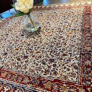May include: A white tablecloth with a floral pattern in shades of red, blue, and yellow. The tablecloth has a fringed edge and is laid on a wooden table. A white flower in a clear glass vase is on the table.