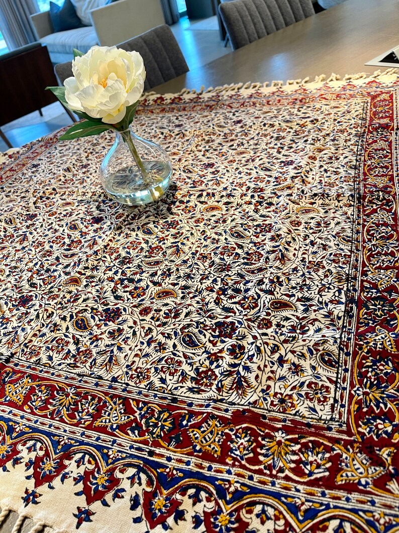 May include: A white tablecloth with a floral pattern in shades of red, blue, and yellow. The tablecloth has a fringed edge and is laid on a wooden table. A white flower in a clear glass vase is on the table.
