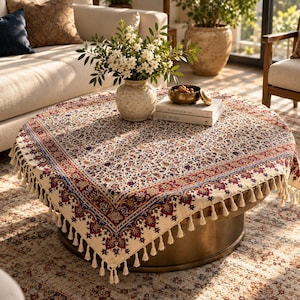 May include: A square tablecloth with a cream-colored base featuring a dense, intricate floral and paisley pattern in shades of red, blue, and gold. The edges are finished with a decorative border and a fringe of small, cream-colored tassels.