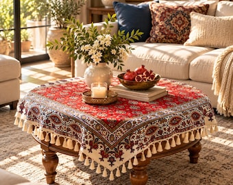 May include: A square table cover featuring a vibrant red center with intricate floral patterns, a wide cream-colored border with paisley motifs, and a decorative fringe trim along the edges, draped over a round wooden table with turned legs.