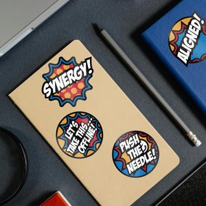 May include: A collection of colorful stickers with bold text and comic-book style designs. The stickers feature phrases like "Synergy!", "Let's Take This Offline!", and "Push the Needle!" on a tan notebook.
